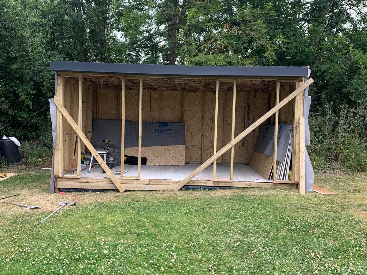 A garden room at frame stage: bare timber studs and bracing under a finished flat roof, open at the front